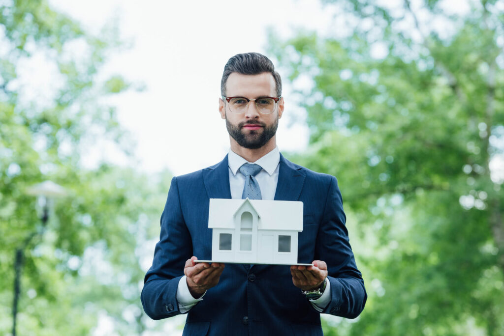 Young businessman holding white house layout and looking at camera while standing in park